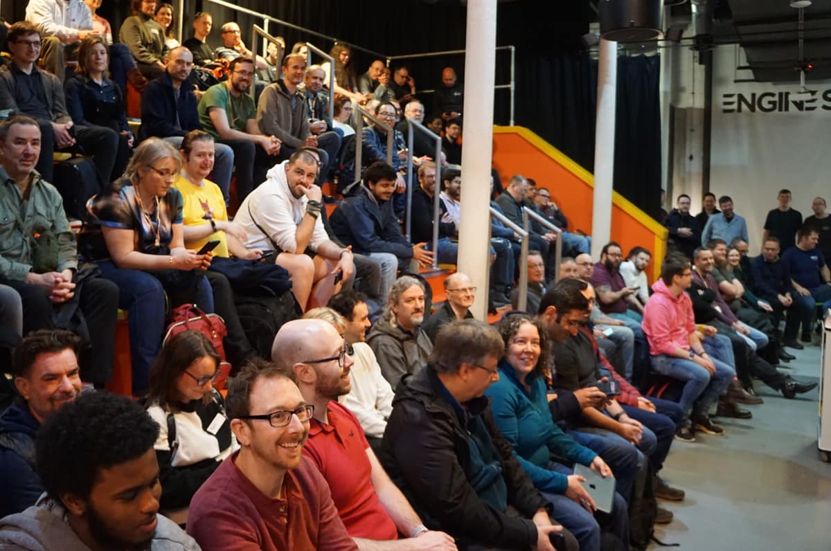 A smiling, full audience on tiered seating waiting for a talk.