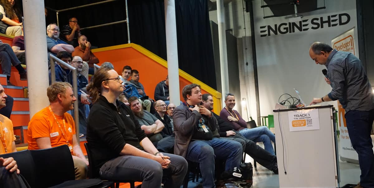 A speaker presenting to the audience in Track 1, with attendees on orange tiered seating.