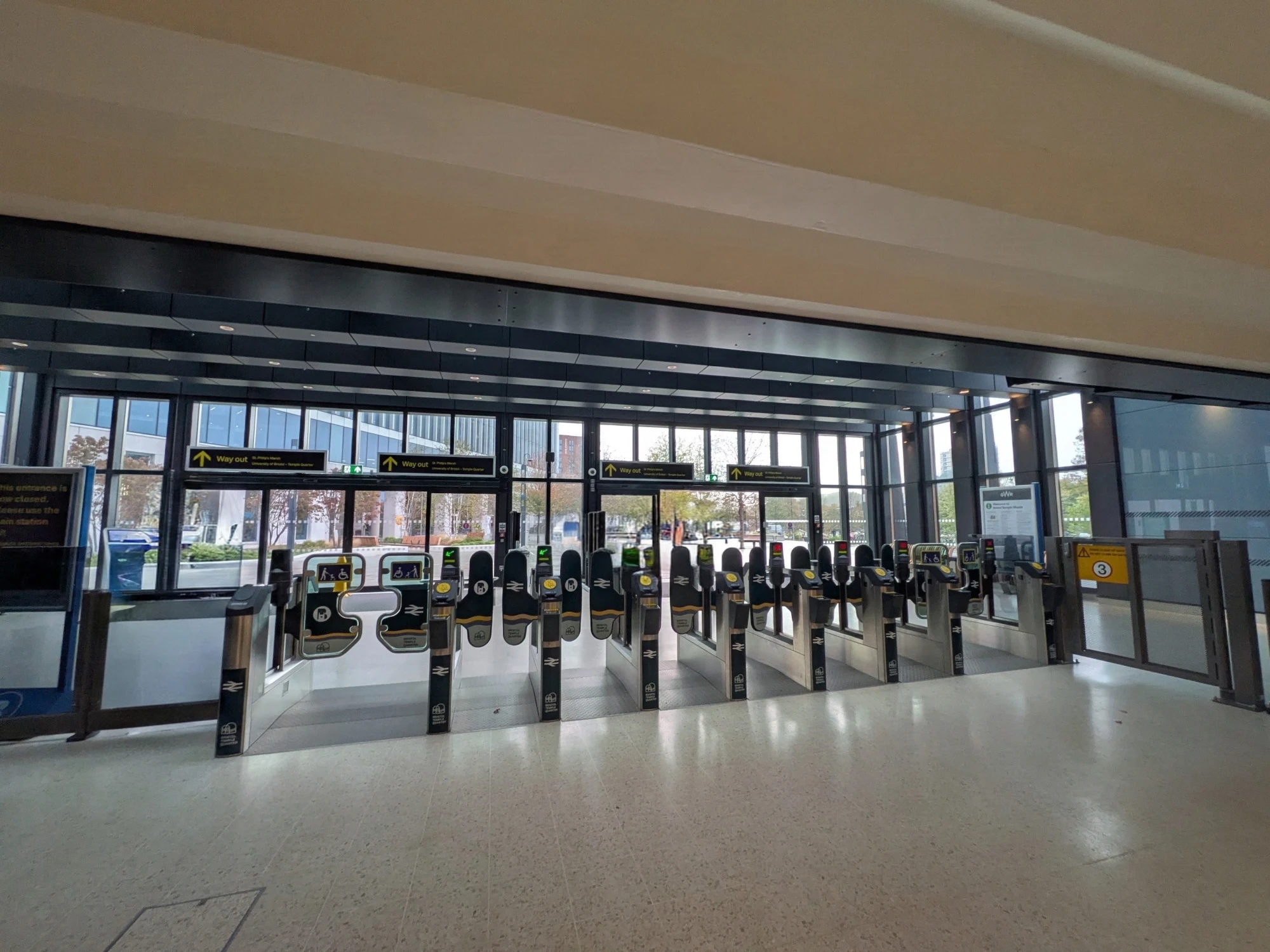Ticket gates at the new exit from Bristol Temple Meads, with 'Way out' signs pointing to the University of Bristol and the campus visible through the glass doors